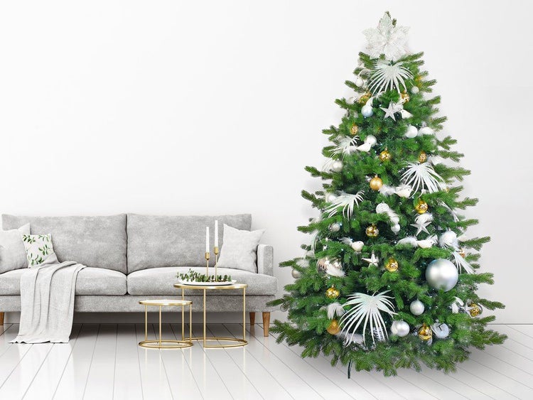 Festlich geschmückter Weihnachtsbaum mit Kugeln und Schmuck in einem Wohnzimmer neben einem grauen Sofa auf hellem Boden.