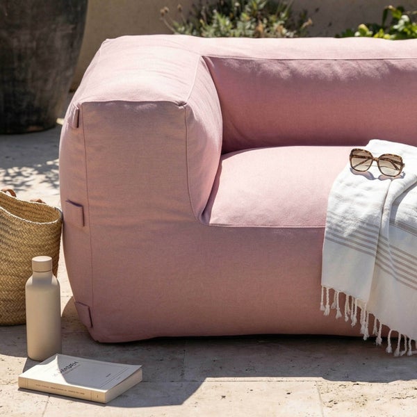 Outdoor-Lounge-Sofa in Pink mit einem Handtuch, Sonnenbrille, Korb, Trinkflasche und Buch auf einer hellen Terrasse.
