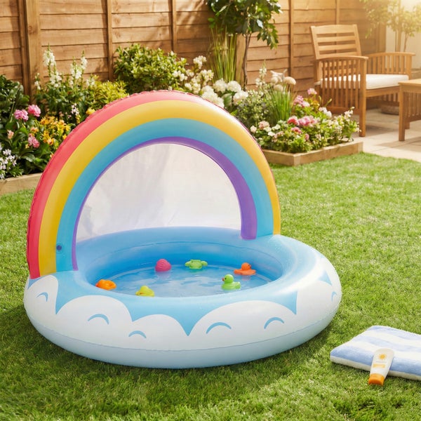 Aufblasbares Kinderplanschbecken mit Regenbogen Sonnenschutz steht auf einer Rasenfläche im Garten neben einem Handtuch und Sonnencreme.