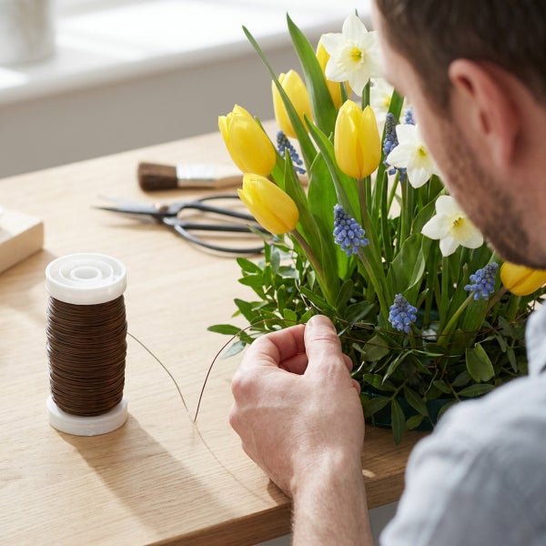 Mann beim Binden eines Frühlingsblumenstraußes mit Wickeldraht auf einem Holztisch mit Schere und Pinsel im Hintergrund.