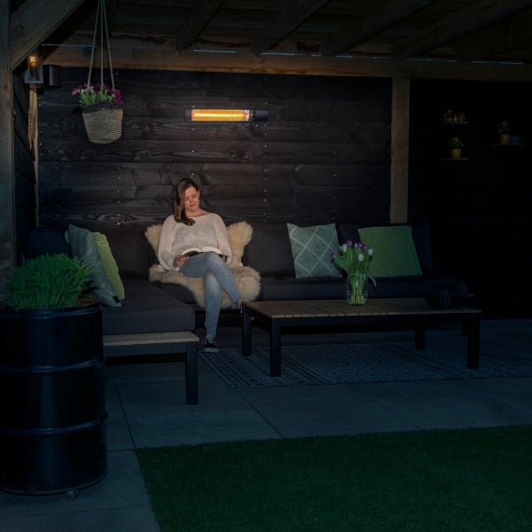Eine Frau liest auf einem grauen Lounge-Sofa unter einer Terrassenüberdachung, beleuchtet von einem Infrarot-Wandheizstrahler an einer Holzwand.
