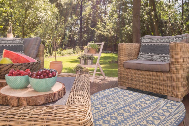 Einrichtung einer sonnigen Holzterrasse mit Rattanmoebeln, Sitzkissen, einem runden Teppich mit Muster und Dekoration mit Obst und Pflanzen.