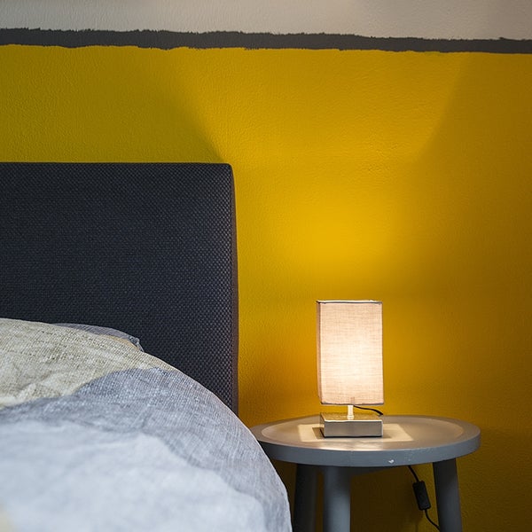 Kleine rechteckige Tischlampe mit Stoffschirm auf einem grauen runden Beistelltisch neben einem Bett vor einer gelben Wand.