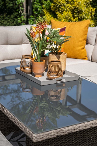 Moderner Garten-Couchtisch aus Glas und Geflecht, darauf ein Tablett mit Pflanzen und Laternen vor einem hellen Gartensofa mit Kissen.