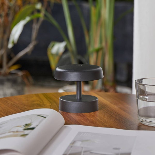 Moderne, mattschwarze tragbare Tischleuchte auf einem Holztisch mit einem Buch und einem Wasserglas.