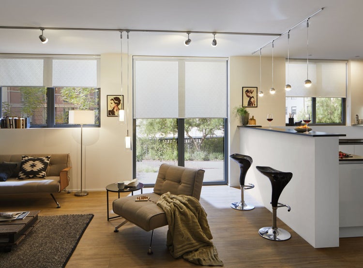 Heller, moderner Wohn- und Küchenbereich mit Schienensystem-Beleuchtung, weißen Rollos, gepolsterten Sesseln und Barhockern auf Holzboden.