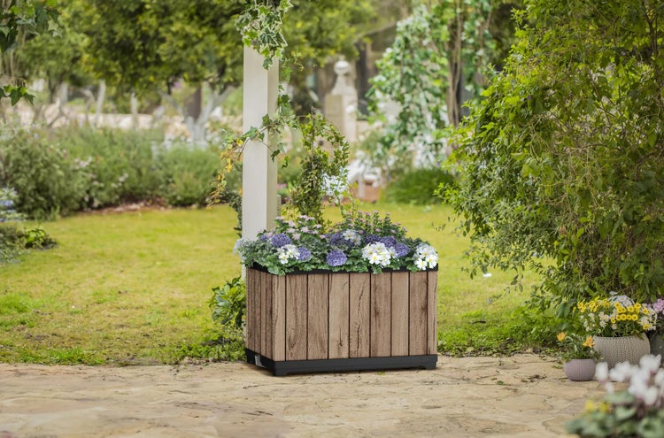 Rechteckiger Pflanzkasten in Holzoptik mit Blumen auf einer hellen Steinterrasse in einem weitläufigen, grünen Garten.