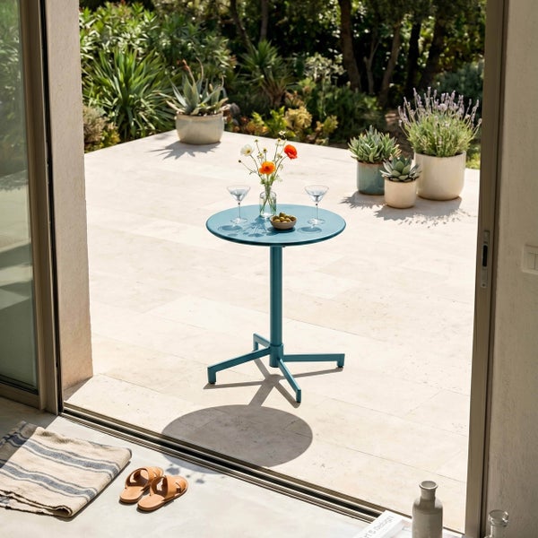 Runder Metall-Gartentisch auf einer hellen Steinterrasse, dekoriert mit Gläsern und Blumen, umgeben von Topfpflanzen und Gartenpflanzen.