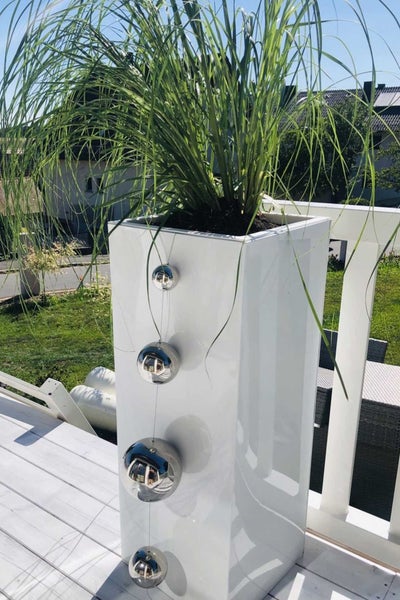 Hoher weißer Pflanzkübel mit grünem Ziergras und dekorativen silbernen Kugeln auf einem hellen Holzbalkon.
