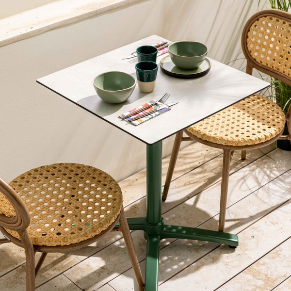 Quadratischer Bistrotisch mit weißer Platte und grünem Gestell, zwei Stühle mit Rattangeflecht und Holzoptik auf einer sonnigen Terrasse.