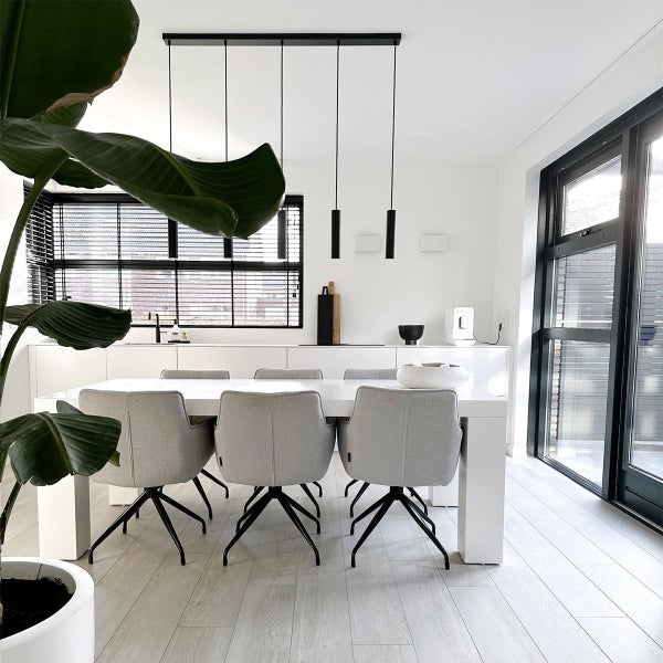 Modernes Esszimmer mit weißem Esstisch und hellgrauen Stühlen mit schwarzem Untergestell unter einer schwarzen Pendelleuchte vor einer Fensterfront.