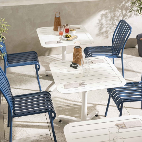 Modernes Garten-Set mit weißen quadratischen Tischen und blauen Metallstühlen auf einer sonnigen Terrasse.