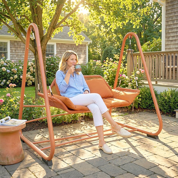 Eine Frau sitzt auf einer modernen Hollywoodschaukel mit Metallgestell und Sitzkissen in einem sonnigen Garten auf einer gepflasterten Terrasse.