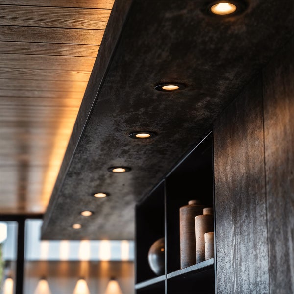 LED Einbaustrahler in einer dunklen Blende mit Steinoptik unter einer Holzlamellendecke mit indirekter Beleuchtung.