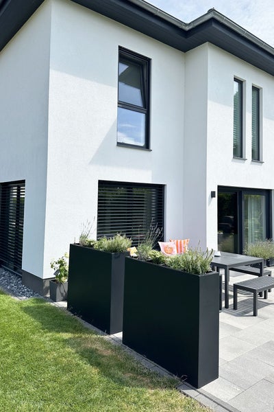 Moderne Terrasse mit großen schwarzen Metall-Pflanzkübeln und einer Sitzgruppe aus Tisch und Bänken vor einem weißen Haus mit dunklen Fenstern.