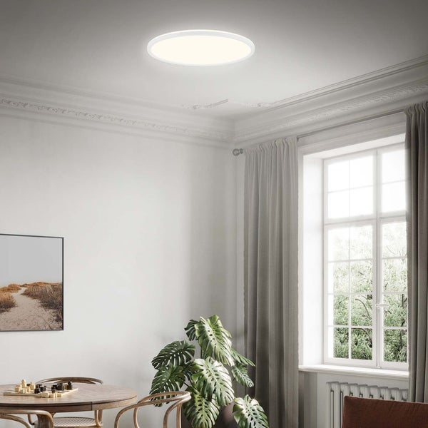 Flaches, rundes LED-Deckenpaneel in einem hellen, modernen Raum mit weißen Wänden, großem Fenster und Holzmöbeln.