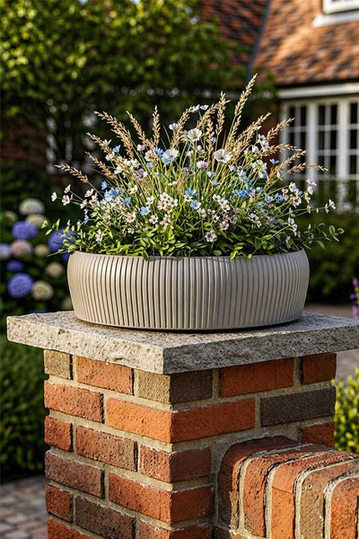 Pflanzschale mit Rillenoptik, bepflanzt mit Feldblumen und Gräsern, platziert auf einem gemauerten Steinpfeiler im Garten.