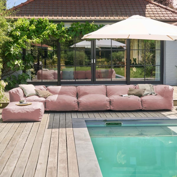Rosa modulares Outdoor-Lounge-Sofa und passender Hocker auf einer Holzterrasse neben einem Pool mit Sonnenschirm und Hausfassade.