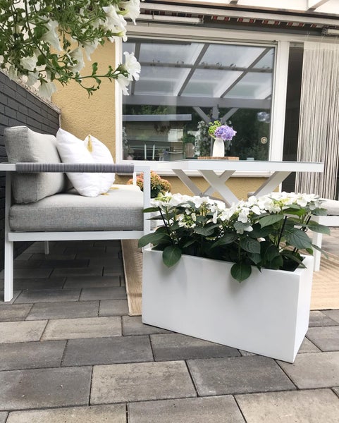 Modern gestaltete Terrasse mit weißem Loungesessel, grauem Polster, weißem Gartentisch und weißem Pflanzkasten mit Hortensien auf Steinpflaster.