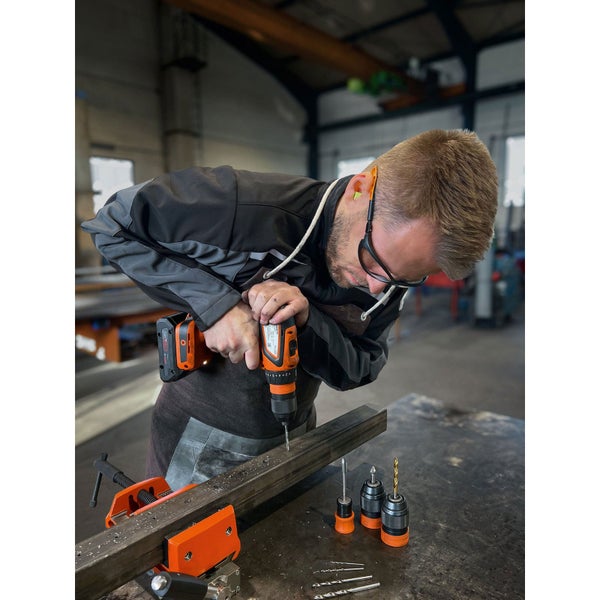 Ein Mann bohrt mit einem Akku-Bohrschrauber in ein Metallstück, wobei er Schutzbrille und Ohrstöpsel trägt. Bohrer und Spannfutter sind ebenfalls sichtbar.