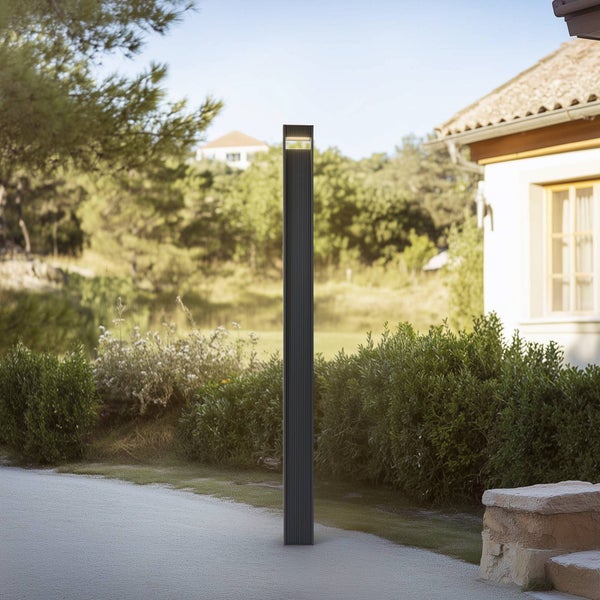 Schlanke, anthrazitfarbene LED-Wegeleuchte in einem modernen Garten neben einem Kiesweg und Sträuchern vor einem Wohnhaus.