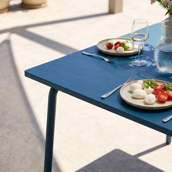 Metall-Gartentisch gedeckt mit Caprese-Salat, Weinglas und Wasserkaraffe auf einer hellen Terrasse im Außenbereich.