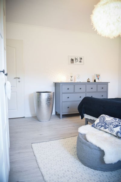 Helles Schlafzimmer mit Boden in Holzoptik, großem silbernem Pflanzgefäß neben grauer Kommode und weißer flauschiger Hängeleuchte.
