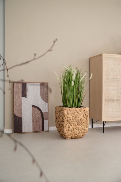 Quadratischer Übertopf aus Korbgeflecht mit hohem Ziergras, ein strukturiertes abstraktes Wandbild und ein Sideboard mit Geflecht.