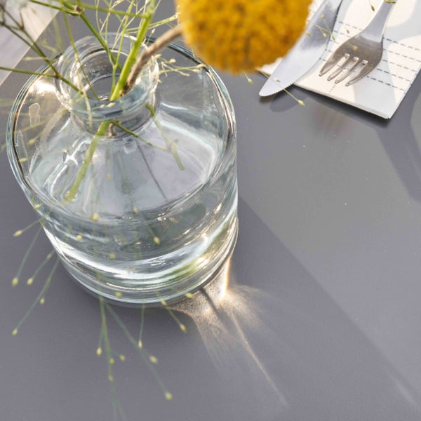 Gedeckter Tisch mit Glasvase, Besteck und Serviette auf grauer Tischoberfläche bei hellem Tageslicht.