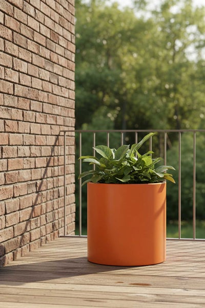 Zylindrischer orangefarbener Pflanzkübel mit grüner Pflanze auf einer Holzterrasse vor einer Ziegelwand.