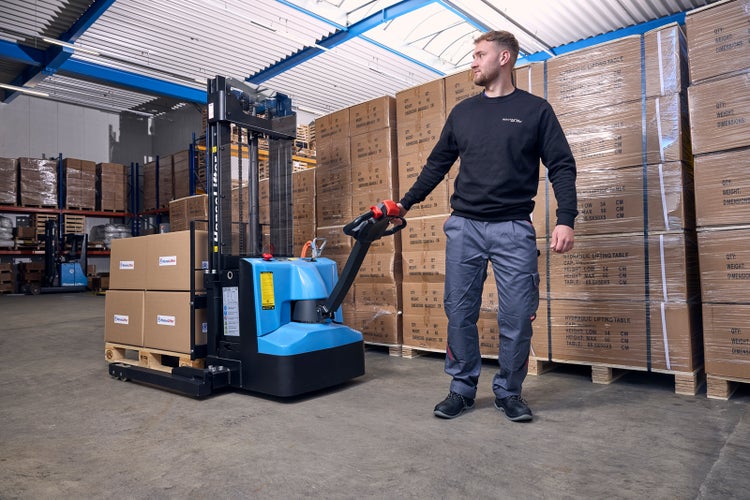 Ein Mitarbeiter bedient einen elektrischen HanseLifter Hochhubwagen in einem hellen Lager mit gestapelten Kartons auf Paletten.