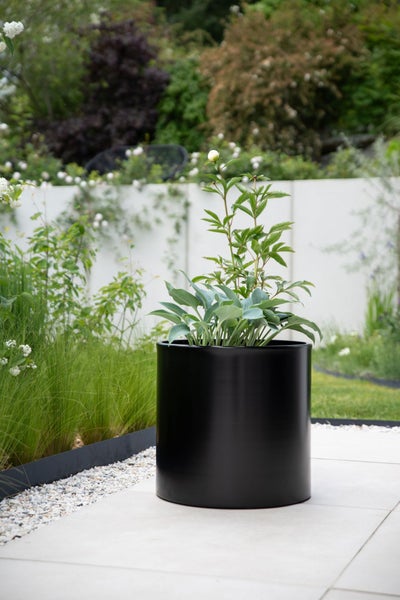 Schwarzer zylindrischer Pflanzkübel mit grünen Pflanzen auf einer modernen Terrasse im Garten.