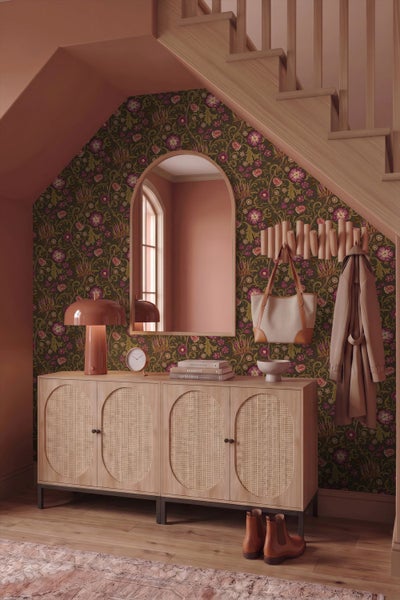 Sideboard mit Rattantüren, florale Tapete, Wandspiegel und Holzgarderobe in einem Flur unter einer Treppe.