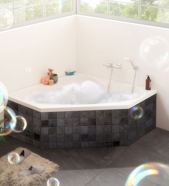Sechseckige Eckbadewanne mit dunkler Fliesenverkleidung und Schaumbad in einem hellen Badezimmer mit Fenster.