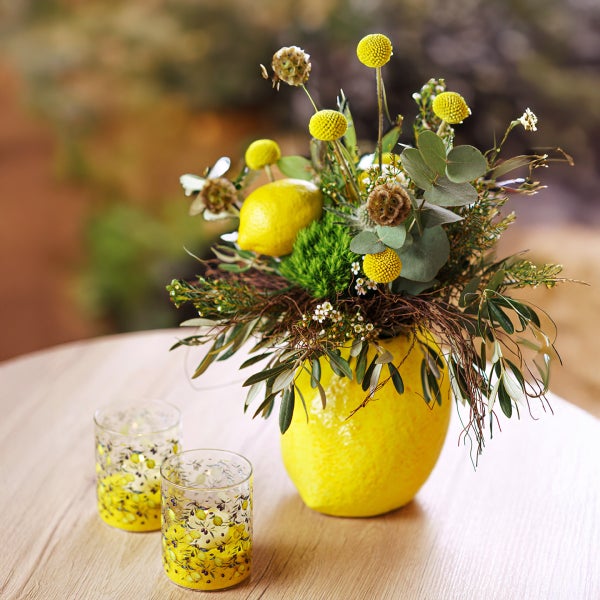 Blumenarrangement mit Zitrone in gelber Vase und Teelichtern auf einem Tisch