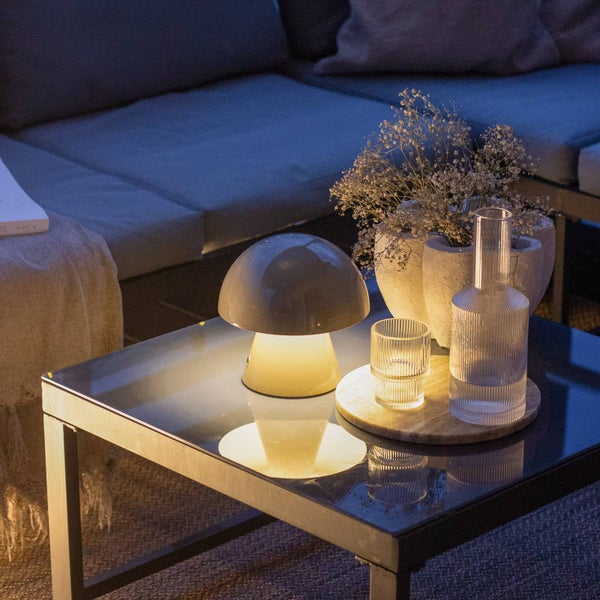 Moderne Tischleuchte in Pilzform auf einem Glastisch in einem gemütlich beleuchteten Loungebereich mit Sofa.