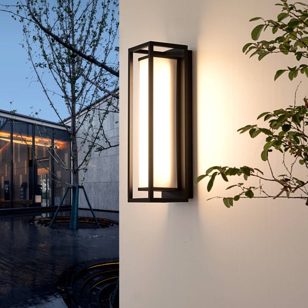 Moderne rechteckige Außenwandleuchte mit schwarzem Metallrahmen und weißem Diffusor, montiert an einer Wand in einer abendlichen Gartenumgebung.