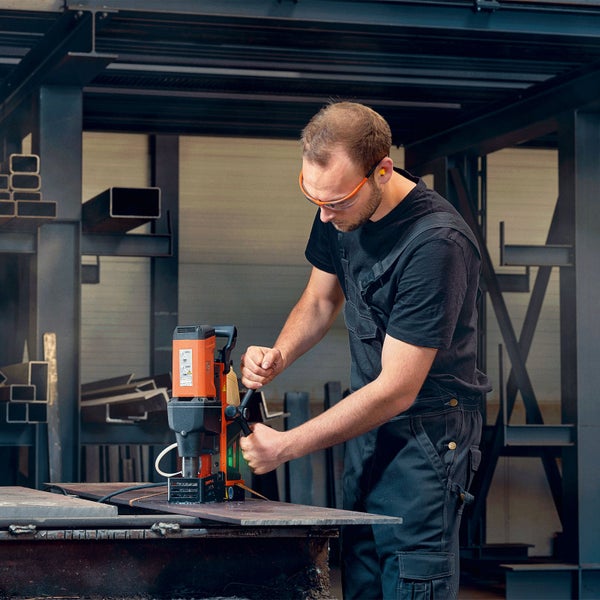 Handwerker mit Magnet-Kernbohrmaschine bearbeitet eine Metallplatte in einer Werkstatt; trägt Schutzbrille und Gehörschutz.