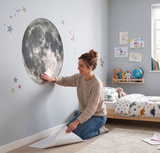 Frau bringt ein großes, realistisches Mond-Wandtattoo in einem hellen Kinderzimmer mit Dinosaurier-Bettwäsche und Sternen-Dekoration an.