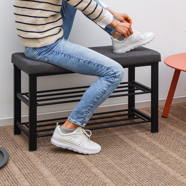 Schuhbank mit schwarzem Metallgestell, grau gepolsterter Sitzfläche und zwei Ablagen für Schuhe, eine Person sitzt darauf.