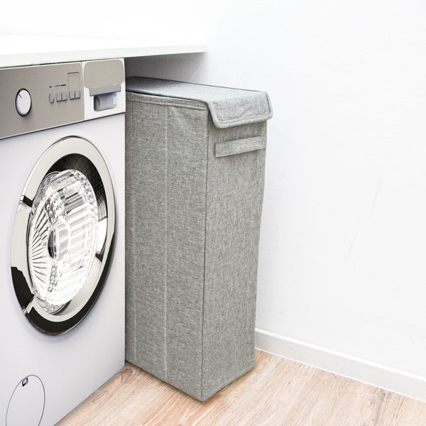 Schmaler Wäschesammler aus grauem Textilgewebe mit Deckel, platziert in einer Lücke neben einer Waschmaschine.
