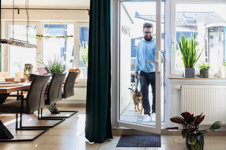 Ein Mann mit Hund betritt durch eine weiße Terrassentür ein helles Wohnzimmer mit Esstisch, grauen Stühlen, Zimmerpflanzen und grünem Vorhang.