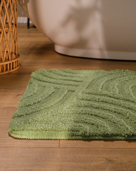 Grüne Badematte mit Wellenstruktur auf Holzfußboden neben einer weißen Badewanne in einem hellen Badezimmer.