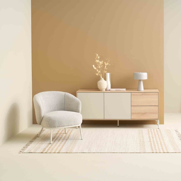 Wohnzimmerszene mit einem texturierten Sessel, einem Sideboard aus Holz mit Türen und Schubladen sowie einem gewebten Teppich mit Fransen.