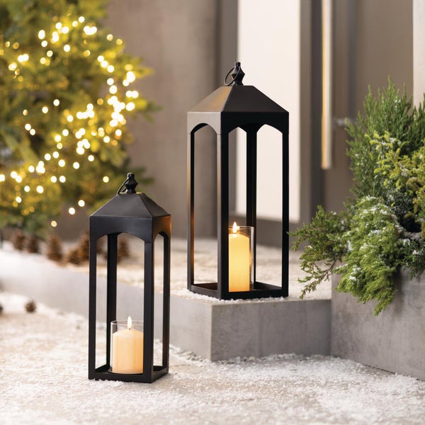 Zwei moderne Metalllaternen mit Kerzen in weihnachtlicher Umgebung mit Lichterkette und Tannen. Dekoratives Licht für den Innen- und Außenbereich.