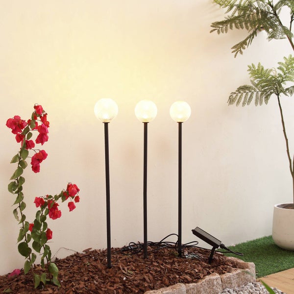 Drei solarbetriebene Kugelleuchten auf schwarzen Stäben in einem Gartenbeet mit Rindenmulch, rosa Blumen und einem externen Solarpanel.