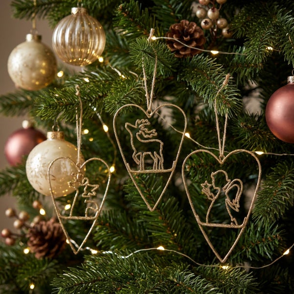 Drei herzförmige Anhänger aus Metall mit weihnachtlichen Motiven an einem Tannenbaum mit Dekokugeln und Lichterkette.