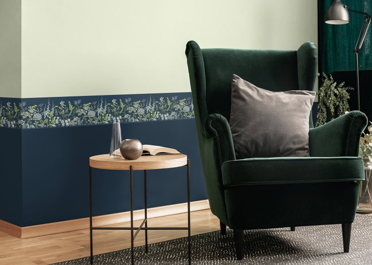 Wohnraumszene mit einer floralen Tapetenbordüre, die eine helle Wand von einer dunkelblauen Wand trennt, neben einem grünen Sessel.