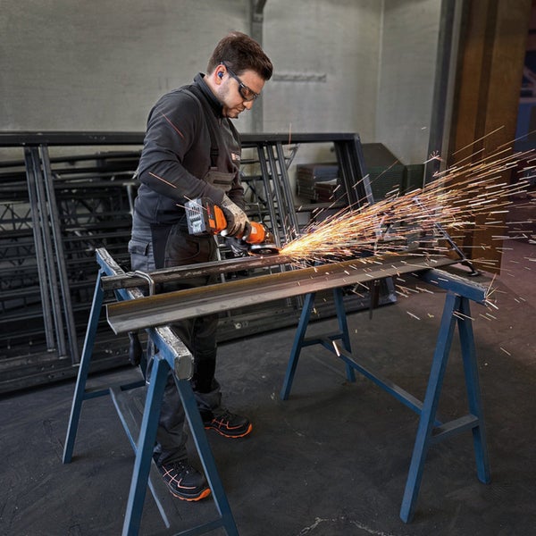 Person in Arbeitskleidung und Handschuhen hält einen Fein Akku-Winkelschleifer und montiert die Schutzhaube in einer Werkstatt.