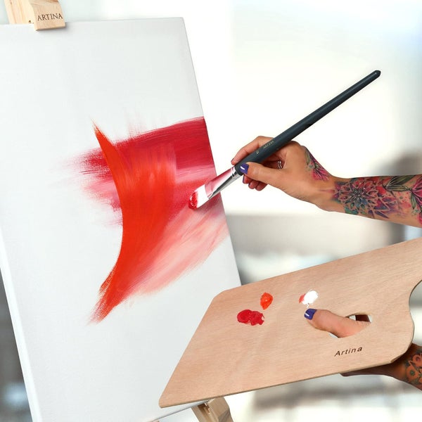 Eine Hand malt mit einem Artina Pinsel auf eine weiße Leinwand und hält eine hölzerne Mischpalette mit roter und weißer Farbe.